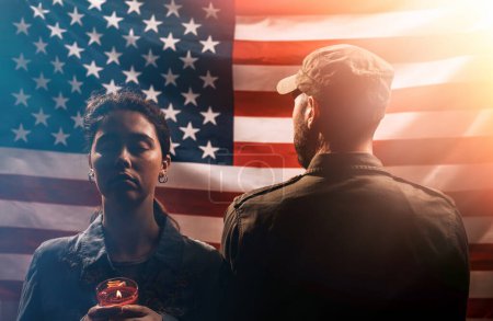 Photo for Veterans day, memorial Day. The soldier stands with his back to a woman holding a lighted candle. Couple on the background of the American flag. The concept of American national holidays and patriotism. - Royalty Free Image