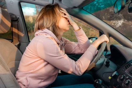 Photo for A young blonde sad woman is sitting behind the wheel of a left-hand drive car and holding her head in despair, covering her eyes with her hand. Side view. Penalty for violation of traffic rules. - Royalty Free Image