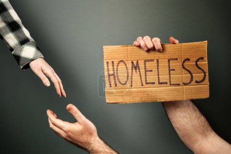 Eine Frau reicht einem Mann eine helfende Hand, der einen Karton mit dem Text Obdachloser in der Hand hält. Dunkler Hintergrund mit grünlichem Farbton. Das Konzept eines sozialen Problems mit Menschen ohne Heimat und Vagabunden.