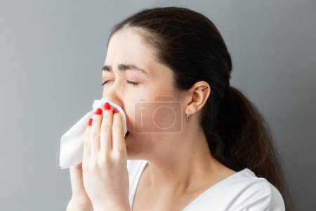 Photo for Portrait of a young Caucasian woman sneezing into a paper handkerchief. Gray background. The concept of allergies, flu and runny nose. - Royalty Free Image