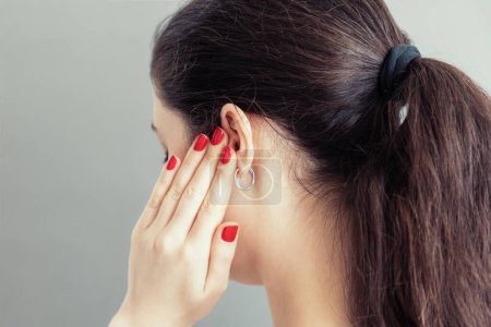 A young caucasian woman closes her ear with her fingers with a red manicure in pain or a loud sound. Gray background. Concept of deafness and ear diseases.