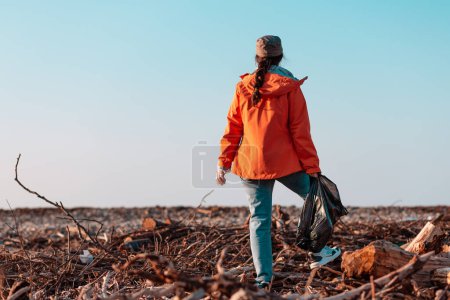 Photo for A young Caucasian female volunteer in a jacket stands with a garbage bag in her hands. In the background, a muddy beach and the sky. Concept of environmental pollution. Copy space. - Royalty Free Image