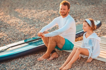 Téléchargez les photos : Père et sa fille assis et se détendre à la plage de galets. Sup board se trouvent près de l'homme. Vacances en famille à la mer. L'été. - en image libre de droit