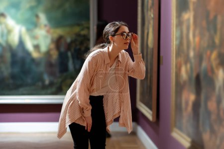 Photo for Side view of young pretty Caucasian woman carefully looking at picture. Masterpieces in background. Concept of the Museum Day. - Royalty Free Image
