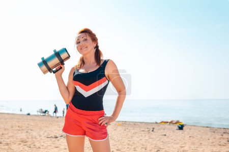 Téléchargez les photos : Sports et entraînement. Jolie femme adulte en tenue de sport, posant sur la plage avec un haltère dans les mains. Espace de copie. - en image libre de droit