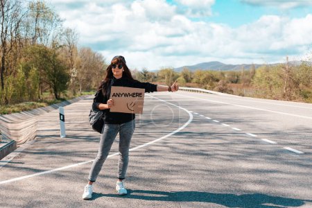 Photo for Smiling young woman in sunglasses and cap holding a cardboard sign with text anywhere. Copy space. The concept of local traveling and hitchhiking. - Royalty Free Image