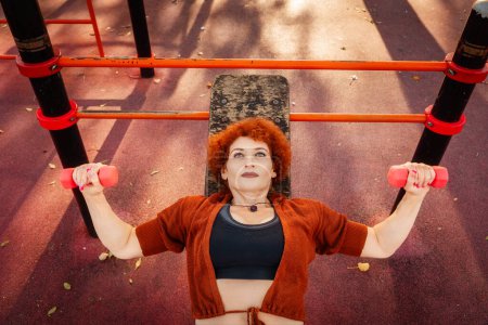 Photo for Top view of Caucasian mid adult woman does french bench press lies on bench training in city park. Copy space. Concept of fitness and sport lifestyle. - Royalty Free Image