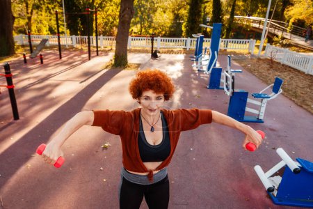 Photo for Top view of mid adult Caucasian woman is doing sports in park on street and looking in camera. Athlete lift up dumbbells in her hands. Healthy lifestyle and sport. - Royalty Free Image