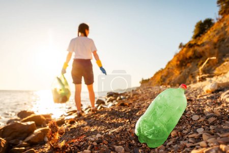 Photo for Defocused back view of woman volunteer in rubber gloves holds full plastic bag of trash. Pollution of pebble wild beach. Concept of protection ocean and Earth Day. - Royalty Free Image
