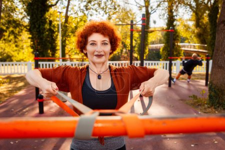 Photo for Training in park. Portrait of adult smiling Caucasian woman looking in camera does stretching with elastic band on sports playground. - Royalty Free Image