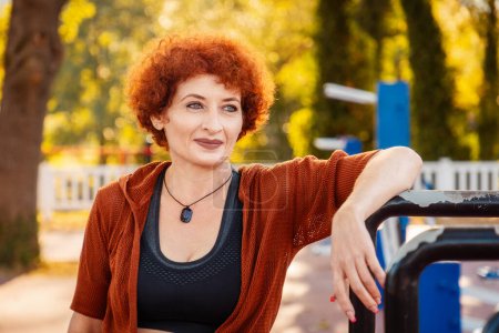 Photo for Portrait of adult Caucasian smiling woman with makeup and ginger curly hair posing in sports park. Concept of healthy lifestyle and fitness. - Royalty Free Image