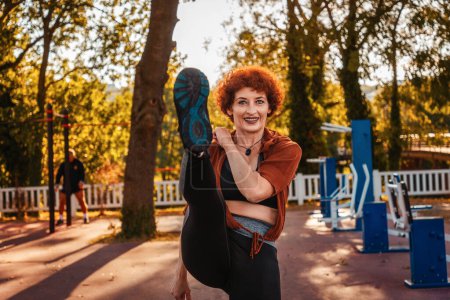 Photo for Happy smiling mid adult Caucasian with ginger hair woman is doing sports lifting leg. Concept of aerobics and cardio training. - Royalty Free Image