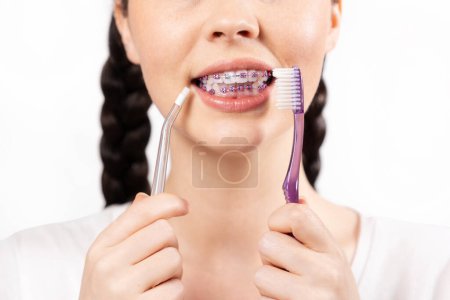 Photo for Close up of young woman with brackets on teeth show manual toothbrush and monopuckle brush. White background. Concept of dental care during orthodontic treatment. - Royalty Free Image