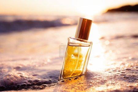 Photo for Close up of rectangular glass bottle with golden perfume washed by sea surf, consecrated by setting sun. Perfumes and cosmetics. - Royalty Free Image