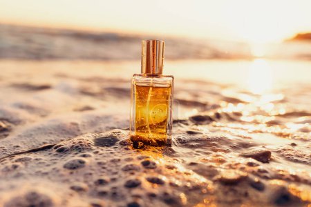 Photo for Close up of rectangular glass bottle with golden perfume washed by sea surf. Sunset light in background. Perfumes and cosmetics template. - Royalty Free Image