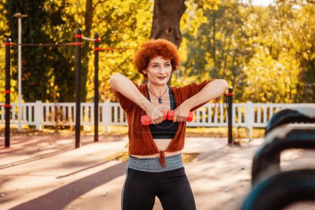 Photo for Mid adult Caucasian ginger hair woman is doing sports in park on street. Athlete's hands lift up dumbbells. Sport lifestyle. - Royalty Free Image