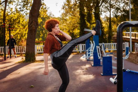 Photo for Fitness and aerobic at city park. Side view of happy smiling mid adult Caucasian woman is doing sports lifting leg. Concept of healthy lifestyle. - Royalty Free Image