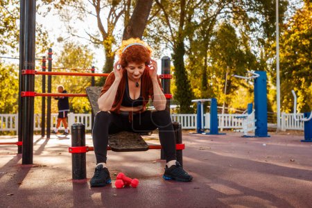 Photo for Mid adult Caucasian woman is sitting on playground and listening to music with headphones. Rest after training. Autumn park in background. Concept of sporty lifestyle. - Royalty Free Image