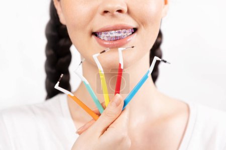 Photo for Close up of young Caucasian smiling woman with brackets on teeth show set of orthodontic toothbrushes. White background. Concept of dental care during orthodontic treatment. - Royalty Free Image