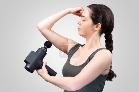 Photo for Side view of adult pretty Caucasian woman gives herself massage on arm with percussion massager-gun with ball nozzle. Gray studio background. Therapy for joint pain relief. - Royalty Free Image