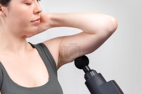 Photo for Close up of Caucasian woman gives herself massage on upper arm with percussion massager-gun with ball nozzle. Gray studio background. Therapy for joint pain relief. - Royalty Free Image