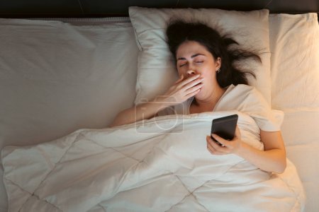Photo for Top view of young Caucasian woman using smartphone and yawning, lying in bed at night. Insomnia, sleepless and social media addiction. - Royalty Free Image