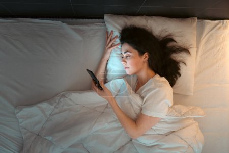 Photo for Top view of young Caucasian woman using smartphone lying in bed at night. Insomnia, sleepless and social media addiction. - Royalty Free Image