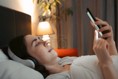 Photo for Close up of young Caucasian woman wearing headphones is lying in bed at night and using smartphone, listening to lullaby music. - Royalty Free Image