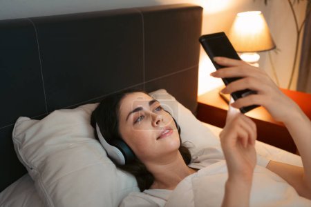 Photo for Young Caucasian woman wearing headphones is lying in bed at night and using smartphone, listening to lullaby music. - Royalty Free Image