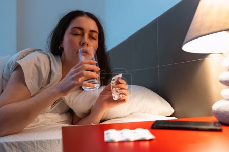Photo for Caucasian adult sleeping woman lies in bed and drink water and holds medicines. Concept of insomnia, sleep disorders and headache. - Royalty Free Image