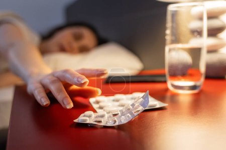 Photo for Defocused woman reaches for red bedside table with medicines, hand and sleeping pills in close-up. Concept of insomnia, sleep disorders and migraines. - Royalty Free Image