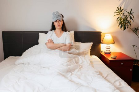 Photo for Disgruntled Caucasian woman wearing sleep mask is sitting in bed with her arms crossed. Insomnia due to noisy neighbors. Copy space. - Royalty Free Image