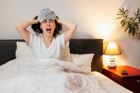 Photo for Irritated Caucasian woman with sleep mask shouts holds hands on head is sitting in bed. Insomnia due to noisy neighbors. - Royalty Free Image