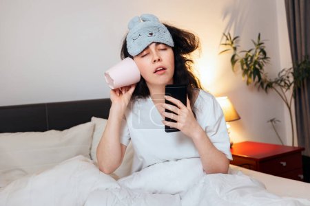 Photo for Portrait of sleeping young woman wearing sleep mask call with cup and drink with phone while sitting in bed. Concept of being late for work and sleepwalking. - Royalty Free Image