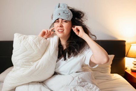 Photo for Portrait of annoyance Caucasian woman wearing sleep mask covers her ears sitting in bed. Insomnia due to noisy neighbors. - Royalty Free Image