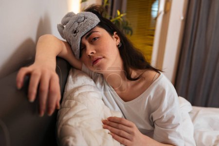 Photo for Tired Caucasian woman wearing sleep mask sitting in bed with pillow. Concept of insomnia and sleepless. - Royalty Free Image