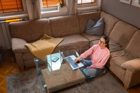 Photo for Top view of young boring Caucasian woman wearing glasses is sitting near sofa and using laptop. Concept of remote work at home office. - Royalty Free Image