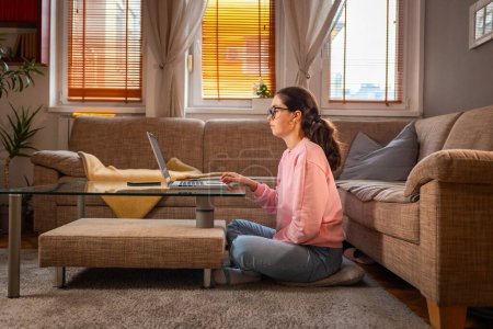 Photo for Side view of young Caucasian woman wearing glasses is sitting near sofa and using laptop. Home office. Concept of remote work and freelance. - Royalty Free Image