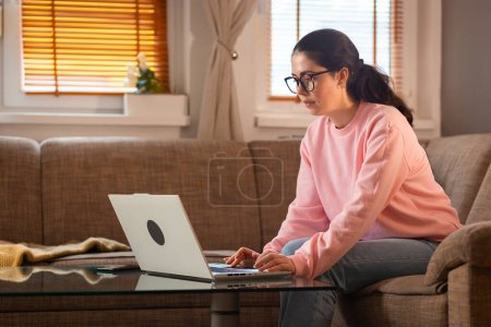 Photo for Young Caucasian woman wearing glasses is sitting on couch and using her laptop. Cozy interior. Concept of remote work and freelancing. - Royalty Free Image
