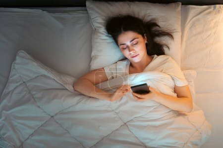 Photo for Top view of young Caucasian woman using smartphone lying in bed at night. Insomnia and social media addiction. Copy space. - Royalty Free Image