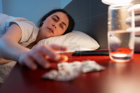 Photo for Caucasian adult woman reaches for red bedside table with medicines. Defocused hand and sleeping pills in close-up. Concept of insomnia, sleep disorders and headache. - Royalty Free Image
