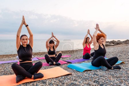 Photo for Outdoor activity at sea coast. Side view of group of Caucasian women meditate sitting on sports mats on pebble beach. Yoga asana for meditating. - Royalty Free Image