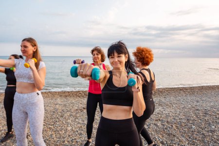 Photo for Outdoor yoga and fitness. Group of happy Caucasian adult fit women are training with dumbbells on pebble beach. Concept of sports lifestyle. - Royalty Free Image