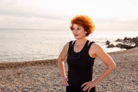 Photo for Outdoor activity. Portrait of adult Caucasian smiling woman with ginger curly hair posing on beach. Copy space. Concept of wellness. - Royalty Free Image