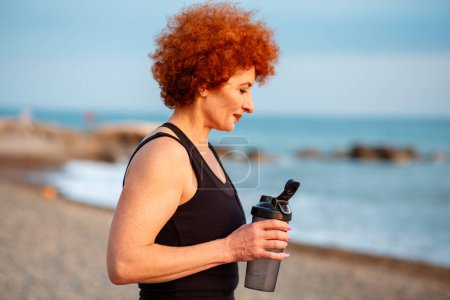 Photo for Side view of Caucasian adult ginger hair woman holds shaker with water. Wellness and healthy lifestyle. - Royalty Free Image