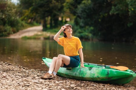 A young happy woman is sitting on a kayak and looking at the sky, holding the visor of her cap. The concept of kayaking and outdoor activities.