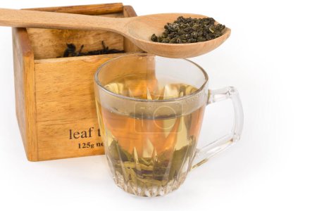 Getrocknete grüne Teeblätter im Holzlöffel und aufgebrühter grüner Tee in der Glasschale vor der hölzernen Teekiste auf weißem Hintergrund