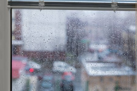Teil des modernen Kunststofffensters mit hochgeklappten Jalousien und Fensterscheibe, die mit Tropfen und Wasserströmen bedeckt ist, und verschwommener Außenumgebung bei Regen, Innenansicht  