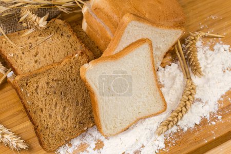 Foto de Pan de trigo blanco en rodajas sin tostar y pan integral para brindar entre las diferentes espigas de cereales en la tabla de cortar esparcida con harina en la mesa rústica - Imagen libre de derechos