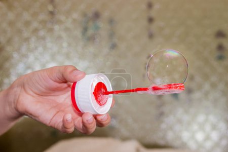 Téléchargez les photos : Iridescent soap bubble on the bubble ring connected by a screw cap of a soapsuds bottle in a child's hand on a blurred background - en image libre de droit